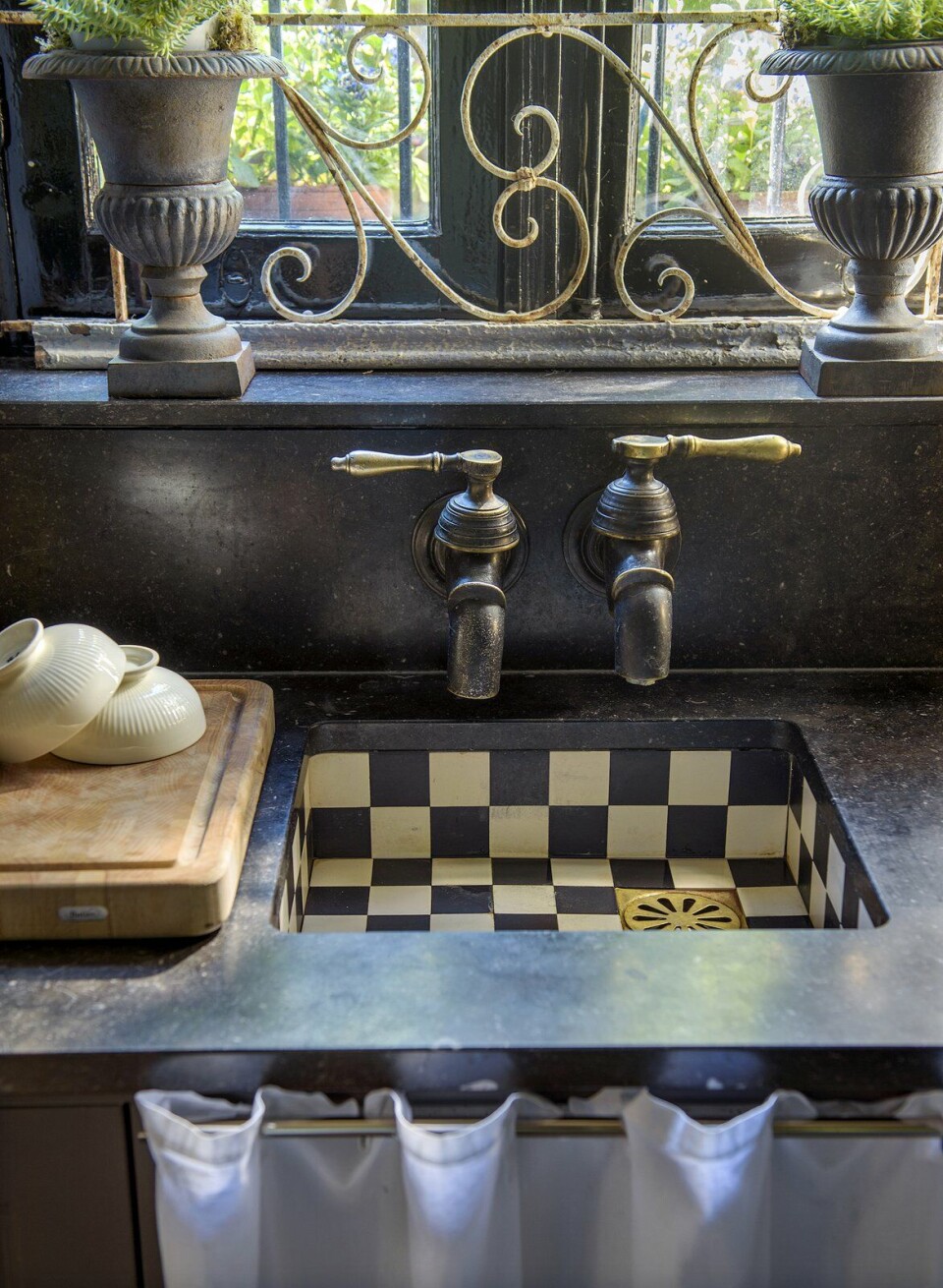 De look-and-feel van een nostalgische boerenkeuken: zwarte en witte tegels, een stoere kraan, paneeldeurtjes, een gordijntje dat de afvoer verbergt en een werkblad van handsteen. binnenkijken aanrecht