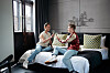 Twee mensen zitten in het art'otel op een bed in een kamer met grote ramen, kleurrijke kussens en een bed aan de muur.