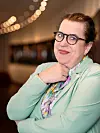 Vrouw met bril in een mintgroene outfit poseert binnenshuis met warme bokeh-verlichting op de achtergrond.