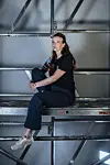 Pride-vrijwilliger Mariska in zwarte kleding zit op een metalen steiger in een industriële ruimte.