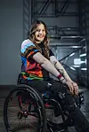 Pride-vrijwilliger Larissa in een rolstoel glimlacht in een industriële ruimte met metalen trappen en constructies.