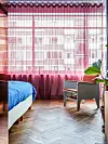 Lichte slaapkamer met blauw beddengoed, fauteuil, planten en grote ramen met rode gordijnen.