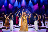 Performer in gouden jurk op een verlicht podium, omringd door dansers in kleurrijke kostuums.