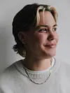 Portret van een vrouw met kort haar, neuspiercing en kettingen tegen een lichte achtergrond.
