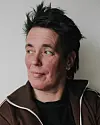 Portret van een vrouw met kort donker haar en een bruine jas tegen een lichte achtergrond.
