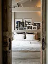 Witte slaapkamer met opgemaakt bed en ingelijste afbeeldingen, gezien door een rustieke houten deur.