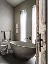 Lichte badkamer met vrijstaand ligbad, wastafel, hoge ramen en een verweerde houten deur.